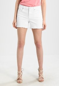 Shorts de mezclilla blanca con dobladillo deshilachado, que cuentan con un diseño de cinco bolsillos, combinados con sandalias de tacón alto de tiras de color beige y una camiseta rosa.