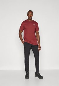 Kratka majica tamnocrvene boje s logotipom, uparena s crnim hlačama i crnim planinarskim cipelama, s uobičajenim krojem i glatkom teksturom.