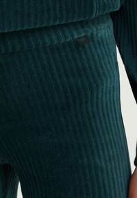 Teal rebrokano blago z navpičnimi črtami, z majhno črno oznako. Mehka tekstura, prileganje oblikovano, primerno za sproščeno nošenje.