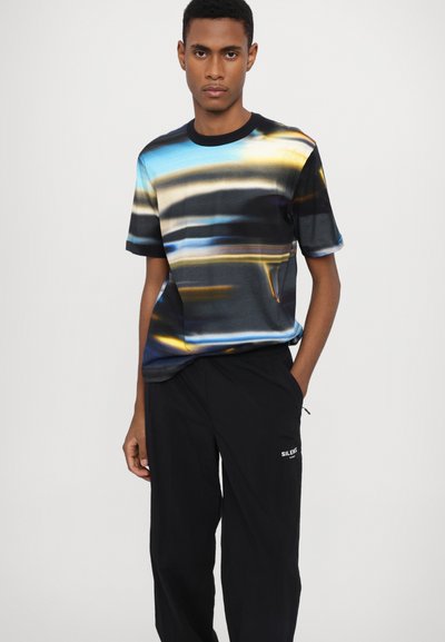 Short-sleeve t-shirt in soft fabric featuring a blue, black, and yellow abstract pattern with a round neckline, paired with black trousers.