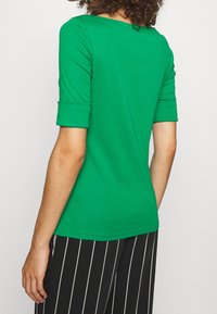 Grön kortärmad topp med rund halsringning, slät textur och uppvikt ärm, i kombination med svart- och vitrandiga byxor.