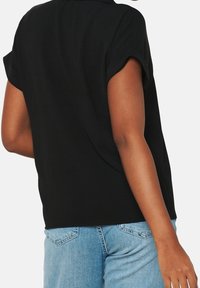 Top corto nero con maniche arrotolate, vestibilità rilassata e tessuto morbido, abbinato a jeans in denim azzurro, mostrato da dietro.