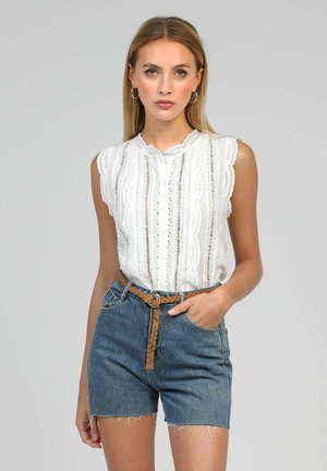 Jeune femme aux cheveux blonds raides portant un chemisier blanc sans manches en tissu ajouré, un short en jean taille haute et une ceinture tressée marron, debout devant un fond gris.