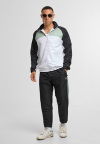 Träningsdräkt med en vit och svart jacka med gröna accenter, som matchas med svarta byxor. Tillverkad av ett mjukt, lätt material.