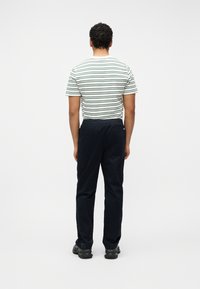 Άνδρας με σγουρά μαλλιά που φοράει λευκό και πράσινο ριγέ μπλουζάκι, σκούρο μπλε παντελόνι και μαύρα παπούτσια, στέκεται κοιτώντας προς ένα απλό λευκό φόντο.