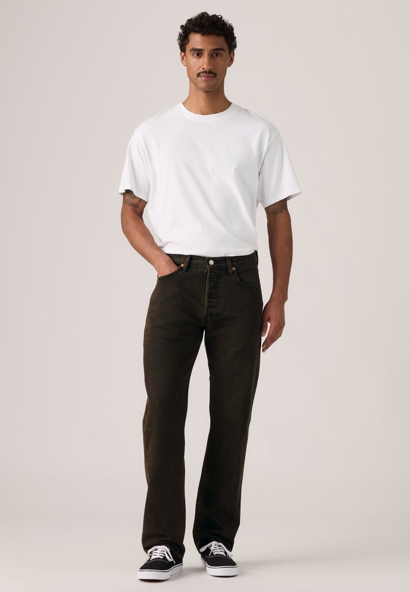 Mand stående med den ene hånd i lommen iført en ensfarvet hvid t-shirt, mørke jeans og sorte og hvide sneakers på en neutral baggrund.