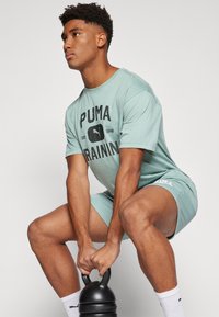 Tricou de antrenament PUMA de culoare verde deschis, cu text negru, asortat cu șorturi, prezintă un design ajustat. Se ține o kettlebell neagră.