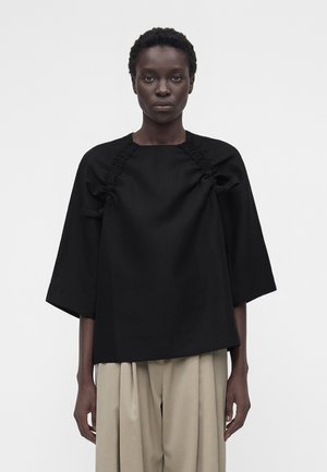 Ženska s kratkimi kodrastimi lasmi nosi ohlapen črn top z naborki in širokimi rokavi, kombiniran z bež plisiranimi hlačami.