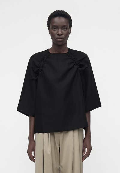 Femme aux cheveux courts et bouclés portant un haut noir ample avec des détails froncés et des manches larges, associé à un pantalon beige plissé.