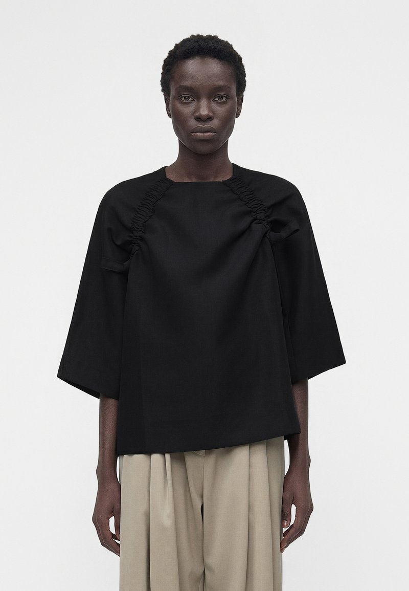 Γυναίκα με κοντά σγουρά μαλλιά φοράει μπλούζα σε χαλαρή γραμμή, μαύρου χρώματος με ρυτιδωτές λεπτομέρειες και φαρδιά μανίκια, συνδυασμένη με μπεζ πλισέ παντελόνια.