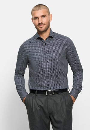 Uomo con capelli corti che indossa una camicia a maniche lunghe con bottoni e fantasia, infilata in pantaloni grigi con cintura nera, mani in tasca, sfondo neutro.