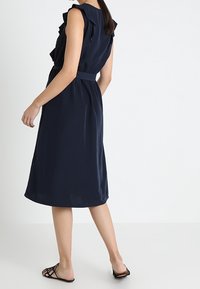 Marinblå midi-klänning med volangdetaljer vid axlarna, åtdragen midja och slät textur. Inkluderar ett matchande bälte och platta svarta sandaler.