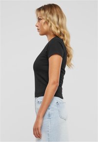 Pikkade blondide juustega naine, kes kannab hästi istuvat musta t-särki ja heledat sinist teksaseelikut, seistes profiilis ühetoonilise tausta ees.