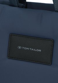 Bolso tote de color azul marino con una etiqueta de cuero negro texturizado. La etiqueta presenta el logo "TOM TAILOR" en plata, cosido de forma segura. Material suave en general.