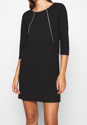 Mujer con un vestido negro hasta la rodilla, con mangas tres cuartos y detalles de ribete blanco a lo largo del frente, posando frente a un fondo liso.