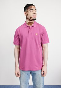 Camisa polo rosa con cuello clásico y mangas cortas, que presenta un pequeño logo amarillo, combinada con vaqueros azul claro.