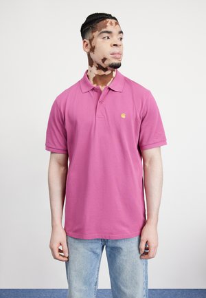 Camisa polo rosa con cuello clásico y mangas cortas, que presenta un pequeño logo amarillo, combinada con vaqueros azul claro.
