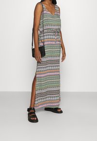 Maxi-jurk met een V-hals, met een meerkleurig patroon van strepen en geometrische vormen, een zijsplit en een getailleerde taille. Zwarte schoudertas en sandalen.