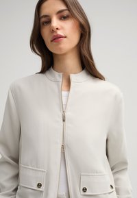 Lichtgrijze zip-up jas met een hoge kraag, twee voorzakken en gouden hardwareaccenten. Glad textuur en minimalistisch ontwerp.