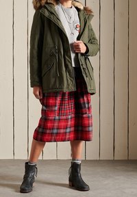 Olive green parka with fur hood, gray graphic sweatshirt, red and black plaid midi skirt, and black ankle boots with wooden heels.