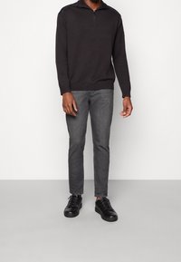 Maglione nero con mezza zip, maniche lunghe e orlo a coste, abbinato a jeans aderenti grigi scuri e sneakers nere, su uno sfondo chiaro.