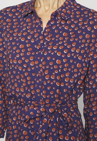 Camicia a maniche lunghe in tessuto viola con pois rossi e bianchi, dotata di colletto, chiusura con bottoni e una cintura legata in vita.