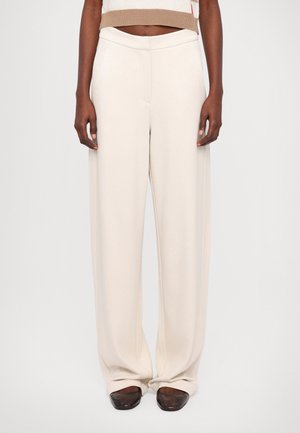 Persoon draagt crèmekleurige broek met hoge taille en brede pijpen met voorzakken en bruine puntige schoenen, staand tegen een effen achtergrond.