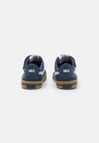 Zilā Nike sporta apavi ar gludu tekstūru, baltām akcentēm un gumijas zoli. Aizmugures skats rāda "NIKE" logotipu uz papēža un mīksto apkakli.