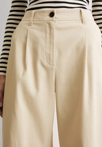 Beige, op maat gemaakte broeken met een hoge taille, voorzien van een enkele voorplooien en een knoopsluiting. Het materiaal lijkt glad met een subtiele structuur.