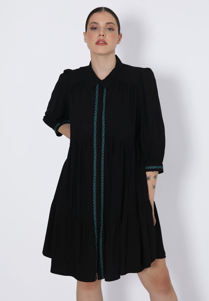 Zwarte jurk met een kraag, opbollende mouwen en een gelaagde rok. Kenmerkt zich door teal geometrische borduur langs het midden en de mouwmanchetten.