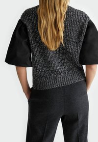 Tlustá pletená vesta tmavě šedé barvy s texturovaným vzorem stehu; má krátké hladké černé rukávy a žebrovaný lem. Černé kalhoty s kapsami.