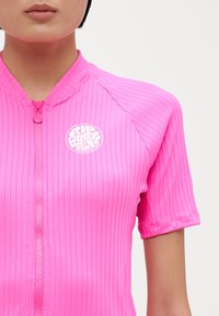 Short-sleeve pink zip-up shirt featuring ribbed texture and a circular white logo on the chest. Zipper extends to collar.