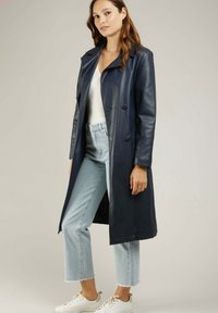 Manteau long en cuir bleu marine avec col cranté, devant double boutonnage et poches latérales. Porté sur un haut blanc et associé à un jean bleu clair.