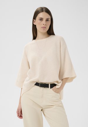 Jeune femme aux cheveux bruns lisses portant un pull beige ample, un pantalon crème avec une ceinture noire et un collier délicat, debout avec la main dans la poche.