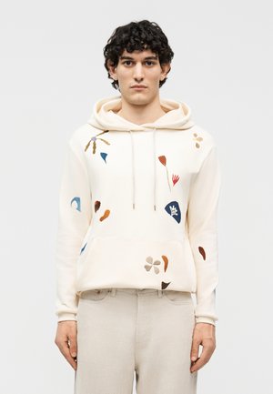 Νέος άνδρας που φοράει εκρού φούτερ με πολύχρωμη αφηρημένη κεντημένη ανθοδέσμη και μπεζ παντελόνι, στέκεται μπροστά σε λευκό φόντο.