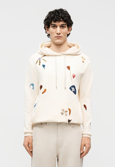 Jeune homme portant un sweat à capuche crème avec une broderie florale abstraite multicolore et un pantalon beige, debout devant un fond blanc uni.