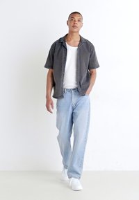 Herrein klær med en grå kortermet skjorte over en hvit t-skjorte, lyseblå rette jeans og hvite joggesko. En enkel bakgrunn.