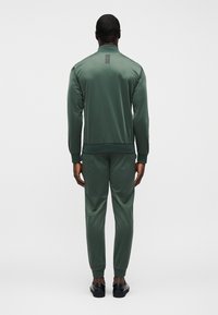 Πράσινη αθλητική φόρμα από λεία ύφανση. Διαθέτει μακριά μανίκια, ψηλό γιακά και ριμπ μανσέτες. Απλό σχέδιο με διακριτική λεπτομέρεια στην πλάτη.