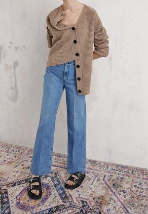 Personne portant un cardigan beige surdimensionné à côtes avec des boutons noirs, un jean large bleu clair, et des sandales noires à boucle, debout sur un tapis à motifs.