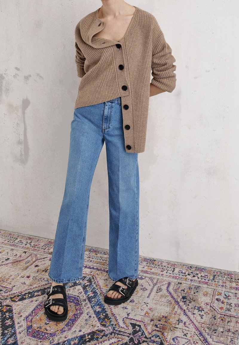 Personne portant un cardigan beige surdimensionné à côtes avec des boutons noirs, un jean large bleu clair, et des sandales noires à boucle, debout sur un tapis à motifs.