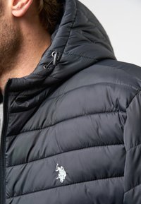 Črna puffer jakna s kapuco, z odebeljenim vzorcem, srebrnim logotipom in sprednjim zadrgo. Gladka tekstura in nastavljiva vezica za kapuco.