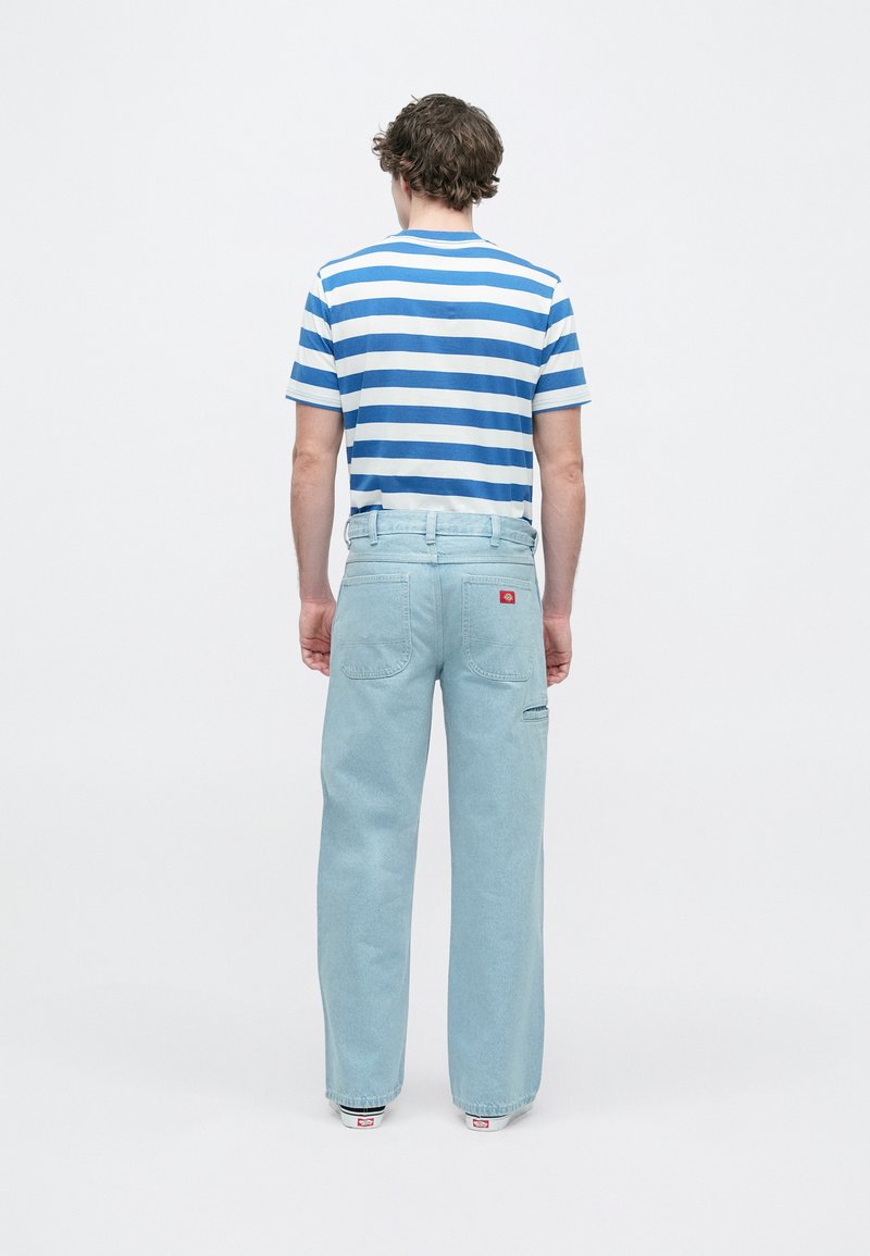 Hellblaue Jeanshose mit lockerem Schnitt, ausgestattet mit Gesäßtaschen und einem roten Logo-Patch. Kombiniert mit einem blau-weiß gestreiften T-Shirt.