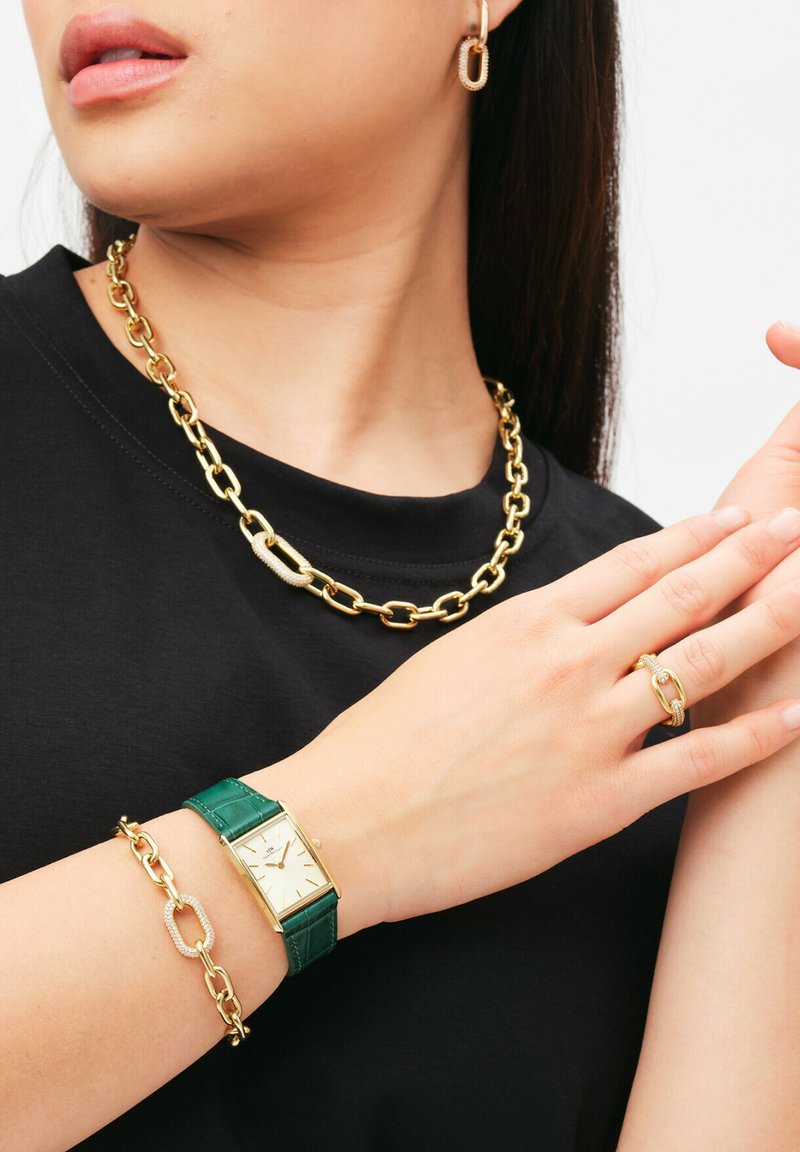 Collana in oro, orologio in pelle verde con accenti dorati e anello e bracciale abbinati. Tutti presentano trame lisce e forme geometriche.