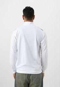 Hvid langærmet polo shirt med en ribbet krave, der har sorte detaljer på ærmerne og en glat, let stofstruktur.