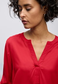 Blusa rossa con scollo a V, realizzata in tessuto leggero e morbido. Presenta un'apertura centrale e maniche leggermente arricciate per un tocco di dettaglio.