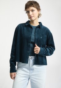Teal kordjacka med krage, knäppning fram och bröstficka; anv�änd över en matchande topp och ljusa denimjeans.