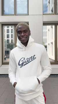 White hoodie with a front pocket, featuring a navy blue "Gant" logo. The model stands with hands in pockets against large windows.