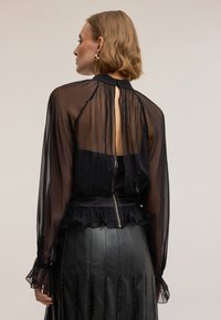 Donna vista di spalle che indossa una camicetta nera trasparente con polsini arricciati e spacco sulla schiena, abbinata a una gonna in pelle nera con dettagli verticali di perline.