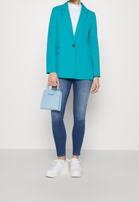 Tyrkysový blazer s jedním knoflíkem, v kombinaci s upnutými modrými džínami a bílými teniskami. Malá světle modrá texturovaná kabelka, stojící ženský model.