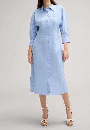 Mujer con un vestido midi de manga larga abotonado y cuello en azul claro con tacones beige de plataforma sobre un fondo liso.